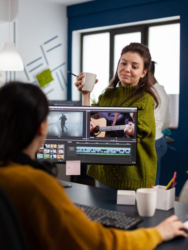 Women videographers editing video project creating content, team of bloggers sitting in modern start up office studio. Manager explaning to colleaque film montage working in post production software.